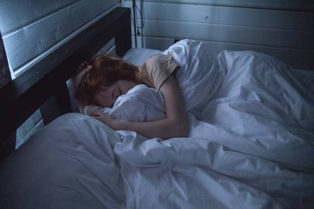 Woman lying on her side in bed, covered with white blankets in a dimly lit bedroom
